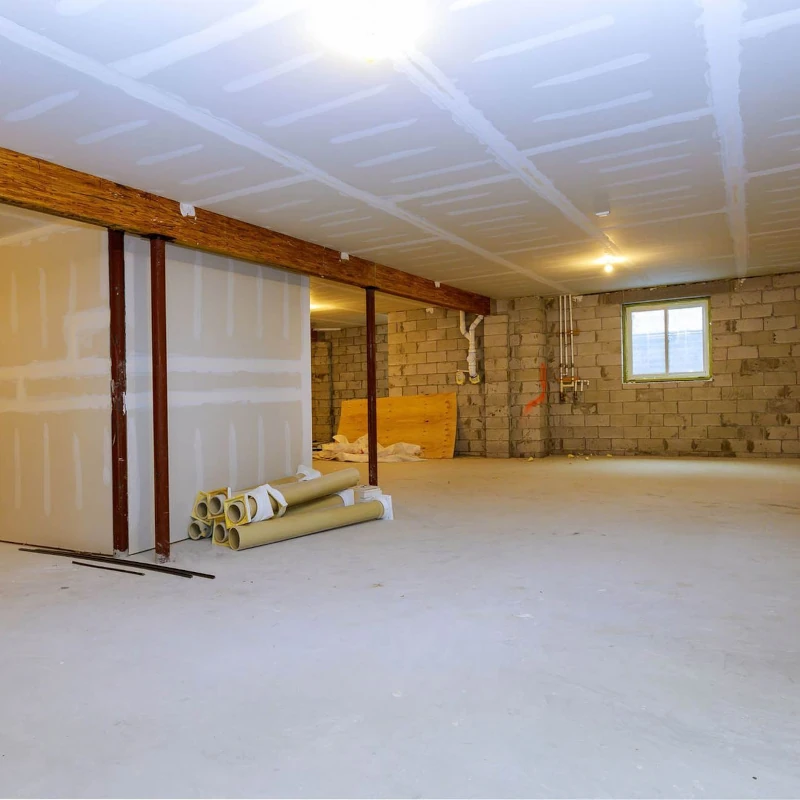 Heller, leerer Kellerraum im Rohbau mit Betonboden, Fenster und Baumaterialien auf dem Boden.